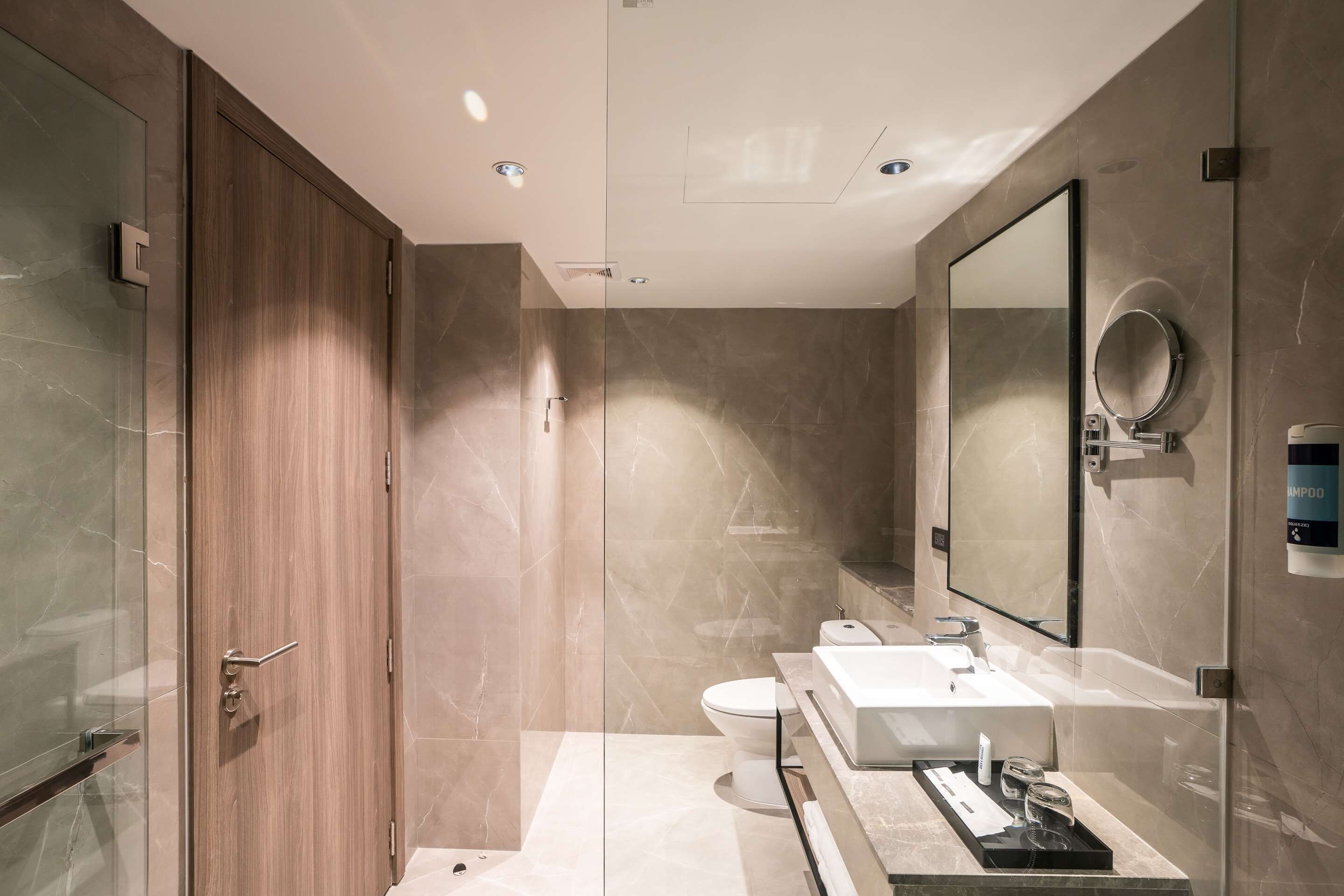 Un baño moderno con cabina de ducha de vidrio, puerta de madera, espejo grande, lavabo blanco y retrete.
