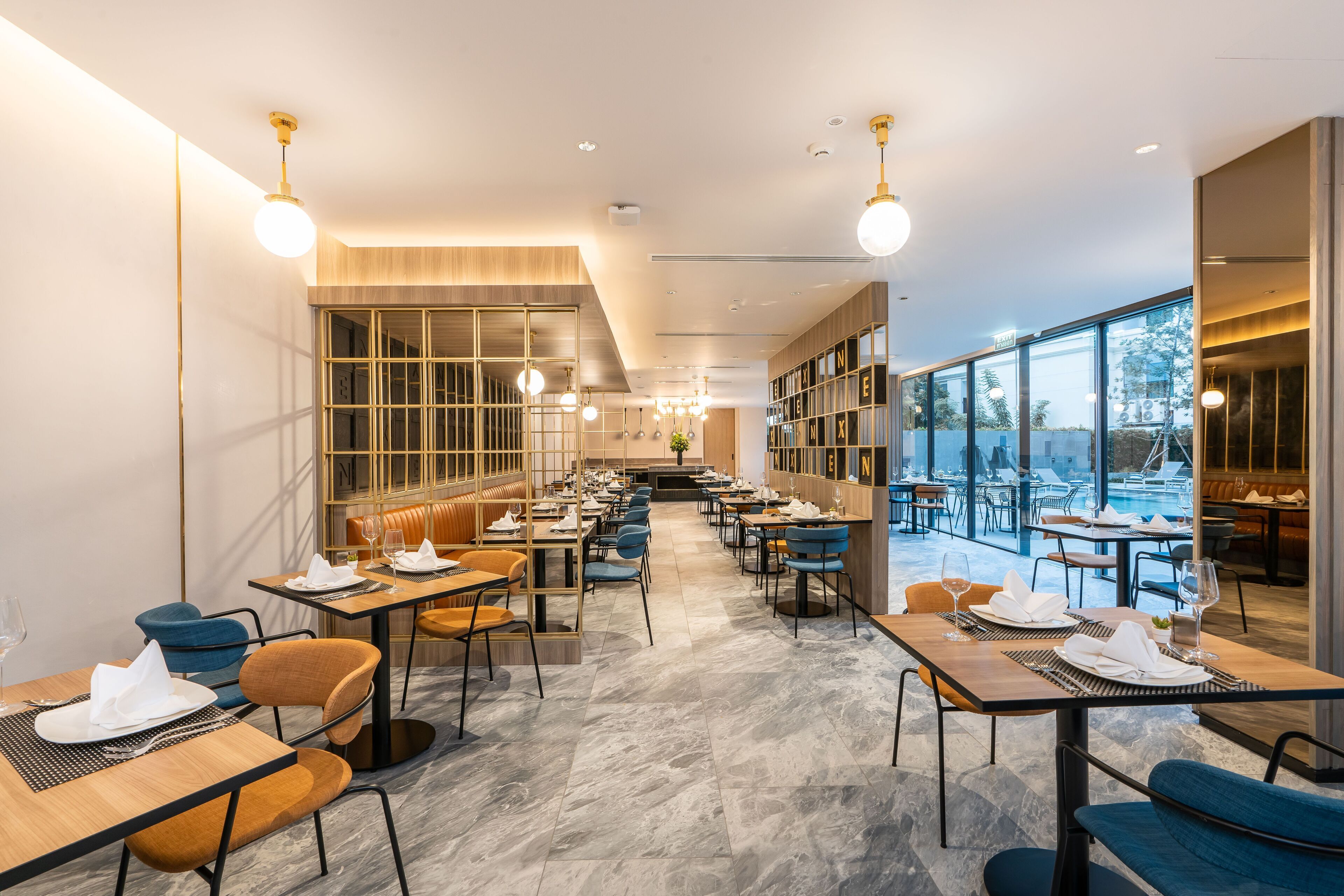 A modern restaurant with a long dining area, high ceilings, and large windows offering a view of the outdoors.