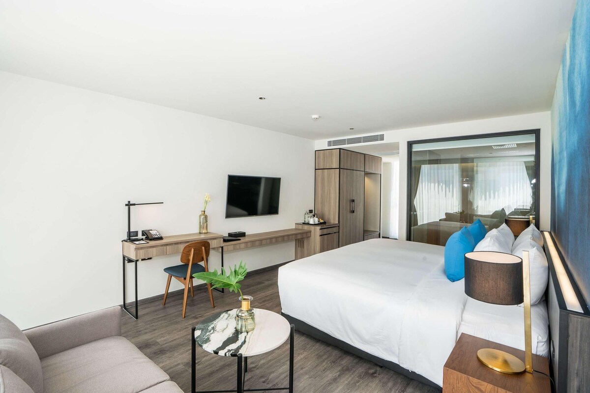 Habitación de hotel moderna con cama, escritorio, silla, televisor y ventana con cortinas.