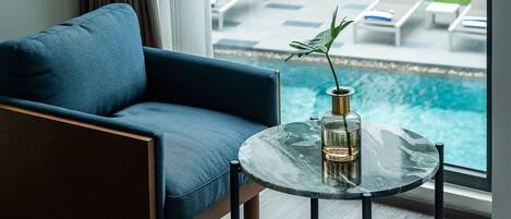 Habitación de hotel moderna con sofá azul, una mesa de centro de vidrio con una planta y vistas a una piscina y jardín.
