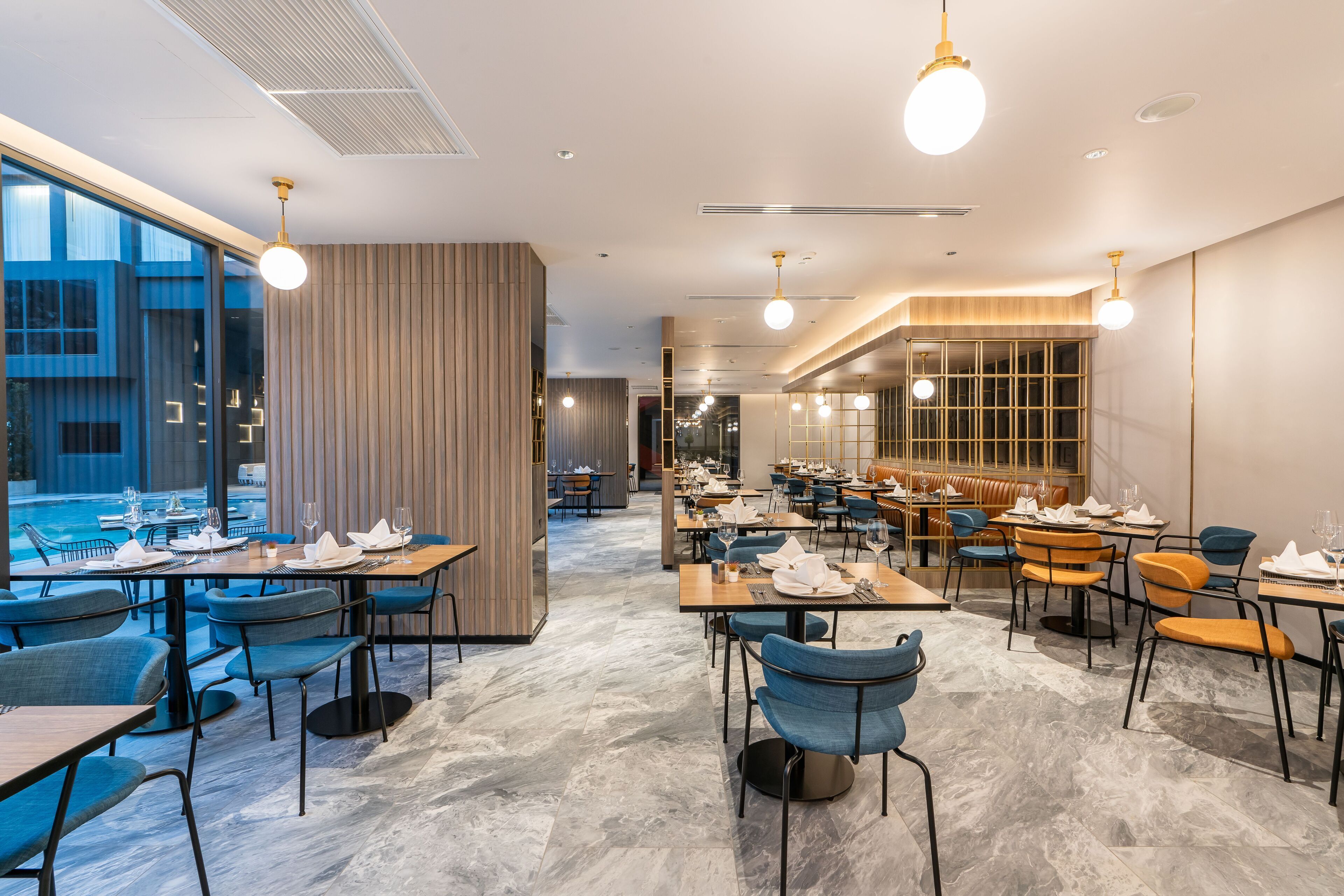 A modern restaurant with a mix of blue and orange chairs, marble flooring, and pendant lighting.
