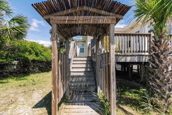 A wooden pathway leading to a covered area with a view of a house and surrounding greenery.