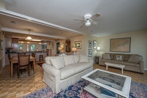 A spacious living area with a beige sofa, a coffee table, and a dining table with chairs. A ceiling fan is visible, and there are framed pictures on the walls.
