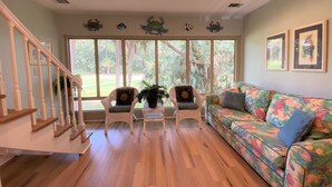 Ein geräumiges Wohnzimmer mit einer Holztreppe, zwei Korbstühlen mit Kissen, einem farbenfrohen Sofa und großen Fenstern mit Blick auf Grünflächen.
