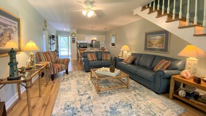 Ein geräumiges Wohnzimmer mit einem blauen Sofa, gestreiften Sesseln, einem Holztisch und einem großen Teppich. Eine Holztreppe führt zu einer oberen Etage.