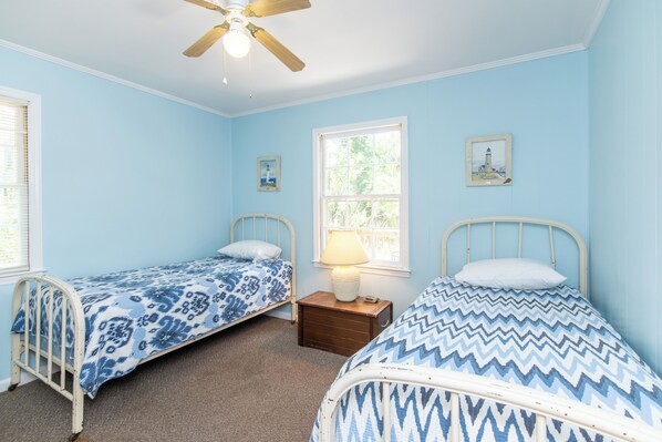 A bedroom with two single beds, a ceiling fan, a window with blinds, and a small bedside table with a lamp.