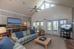 Un salon spacieux avec un plafond haut, un ventilateur de plafond, un grand téléviseur à écran plat, une bibliothèque, un canapé et une table basse.