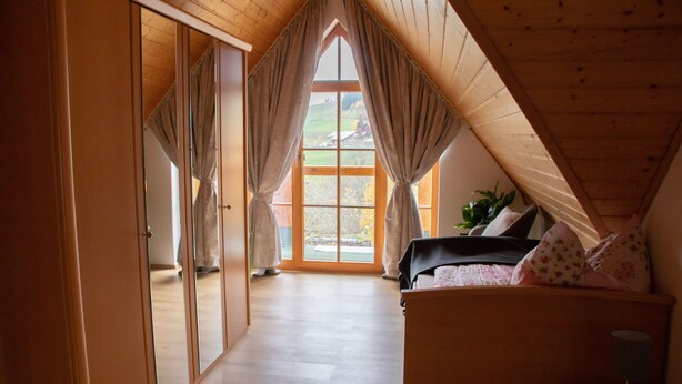 A bedroom with a wooden ceiling, a bed with floral bedding, and a view of a green landscape through a large window. - Ferienhaus Rudenberg (Titisee-Neustadt)