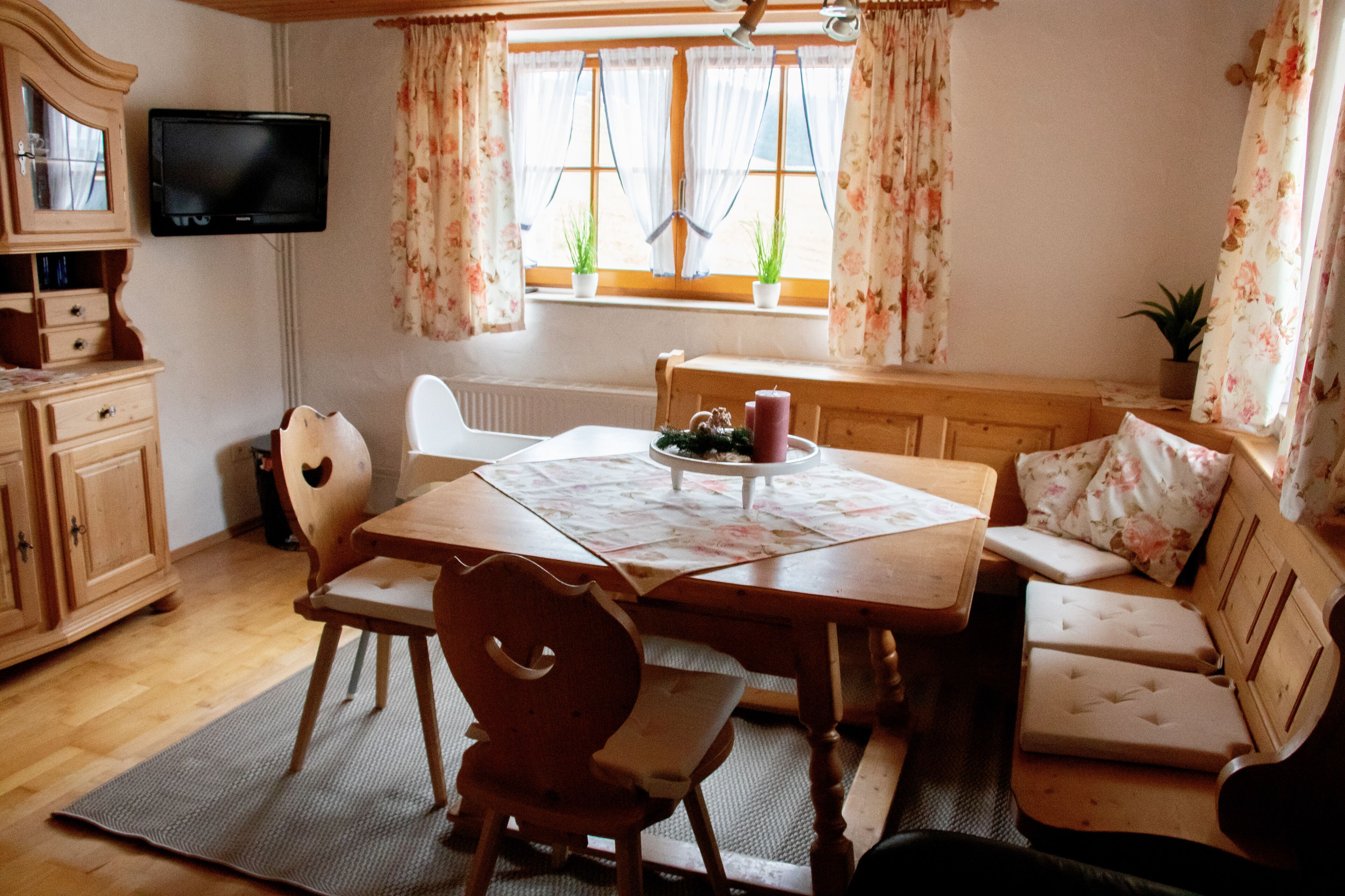 A cozy dining area with a wooden table, chairs, and a bench. A television is mounted on the wall, and there are windows with curtains.