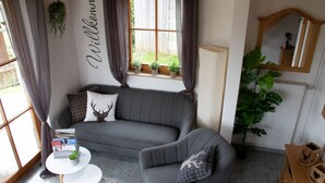 A cozy living room with a grey sofa, a small white side table, and a large window with curtains. - Ferienhaus Rudenberg (Titisee-Neustadt)