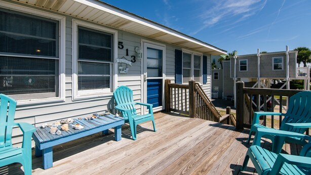 A wooden deck with blue Adirondack chairs and a table, overlooking a building with large windows and a number '513' painted on the wall.