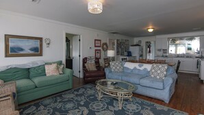 A living room with a green sofa, a blue sofa, a glass coffee table, and a patterned rug. There is a kitchen in the background with a large window.