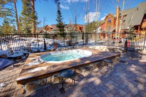 A hot tub on a stone patio with a wooden deck, surrounded by a metal fence, with houses in the background. - Seasons 1825 (Keystone)