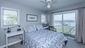 A bedroom with a bed, a desk, a chair, a ceiling fan, and a large window with a view of the water.