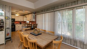 Un comedor con una mesa y sillas de madera, una cocina al fondo y ventanas grandes con cortinas.