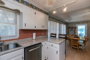 A kitchen with white cabinets, a granite countertop, a dishwasher, and a dining area with wooden chairs and a table. - Pet Friendly w/ Fenced Yard; Screened Porch & Sun Deck; Easy Beach Access; Cable TV/WiFi (Edisto Beach)