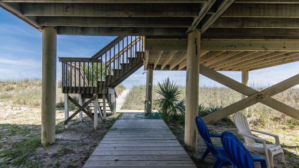 Une terrasse en bois avec un escalier menant à une zone abritée, entourée de sable et d’une végétation clairsemée.