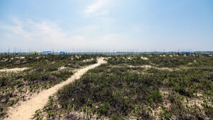 Um caminho de areia serpenteia por uma área gramada com vegetação escassa e céu claro.