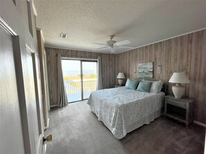 A bedroom with a large bed, two bedside lamps, a ceiling fan, and a view of the sea through a sliding door.