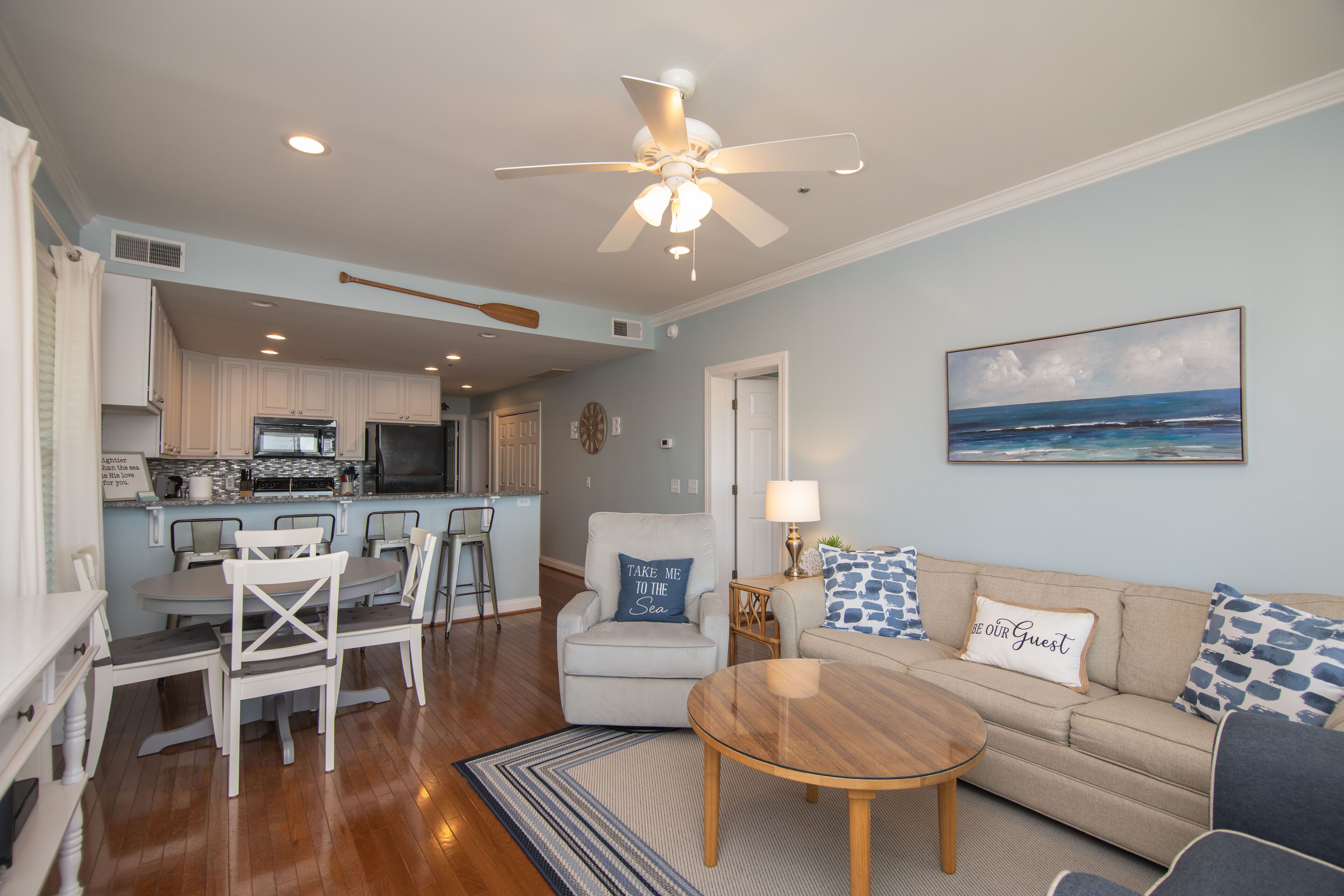 A spacious living room with a round wooden coffee table, a beige sofa with patterned cushions, and a dining area with white chairs and a kitchen in the background.