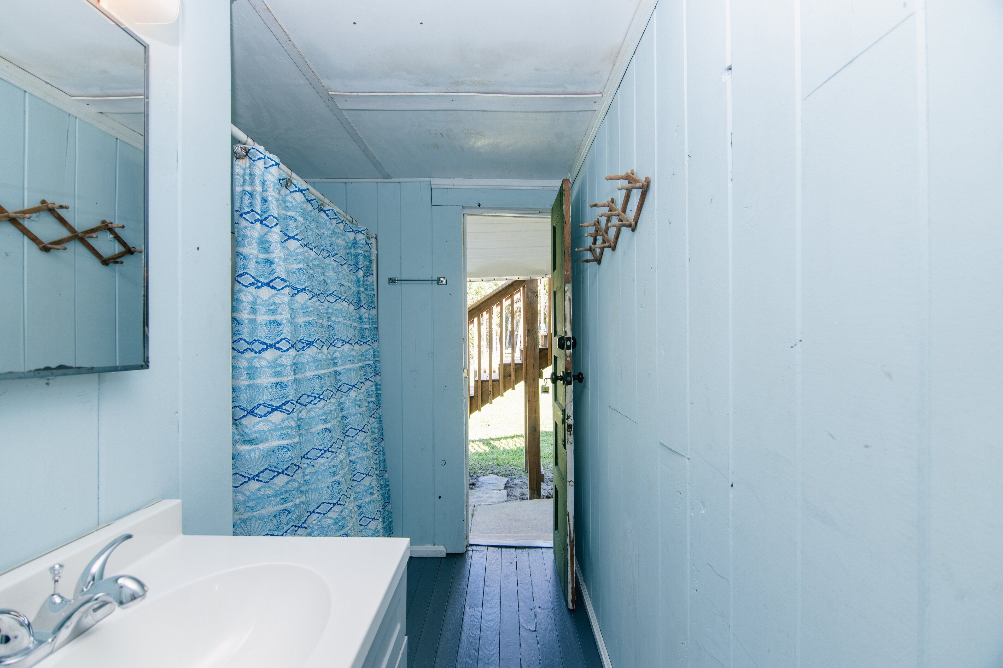 Un baño estrecho con un lavamanos blanco, cortina de ducha de motivos azules y una puerta de madera que da a un espacio al aire libre.