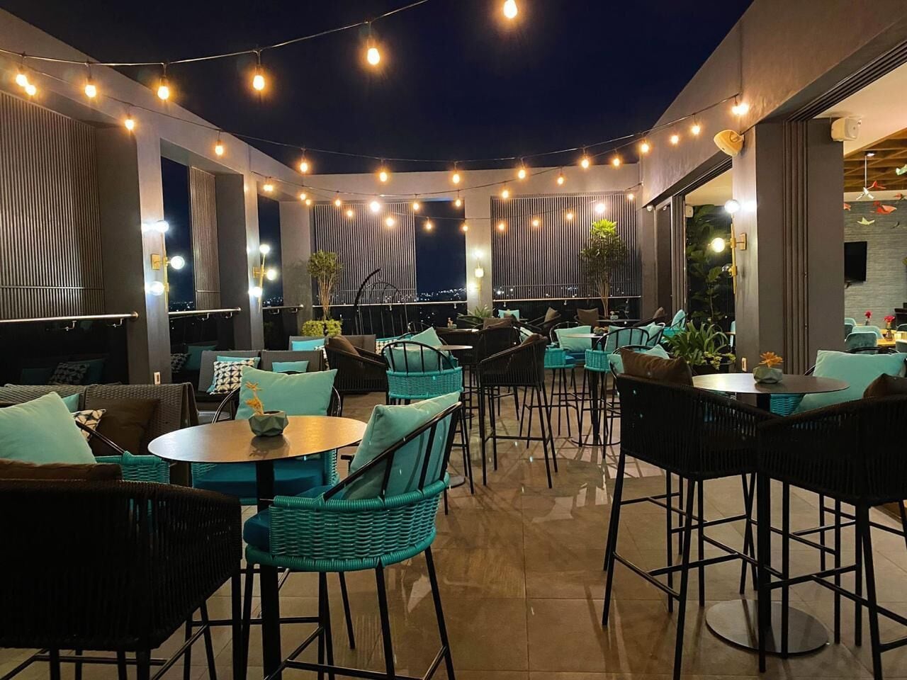 Ein Terrassenbereich im Freien mit runden Tischen und Stühlen, Lichterketten und Kübelpflanzen.