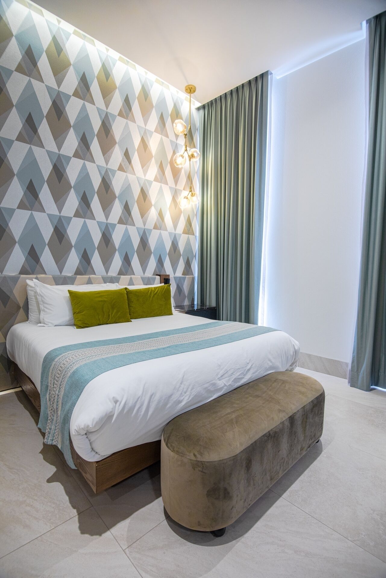 Um quarto moderno com parede estampada em padrão geométrico, uma cama grande com travesseiro verde, um banquinho e um luminário pendente.
