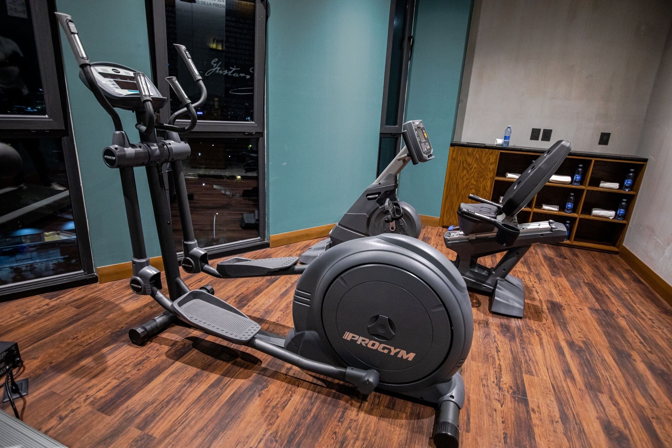 Een sportschool met een Progym elliptische machine, een gewichtbank en een wandrek met handdoeken en waterflessen.
