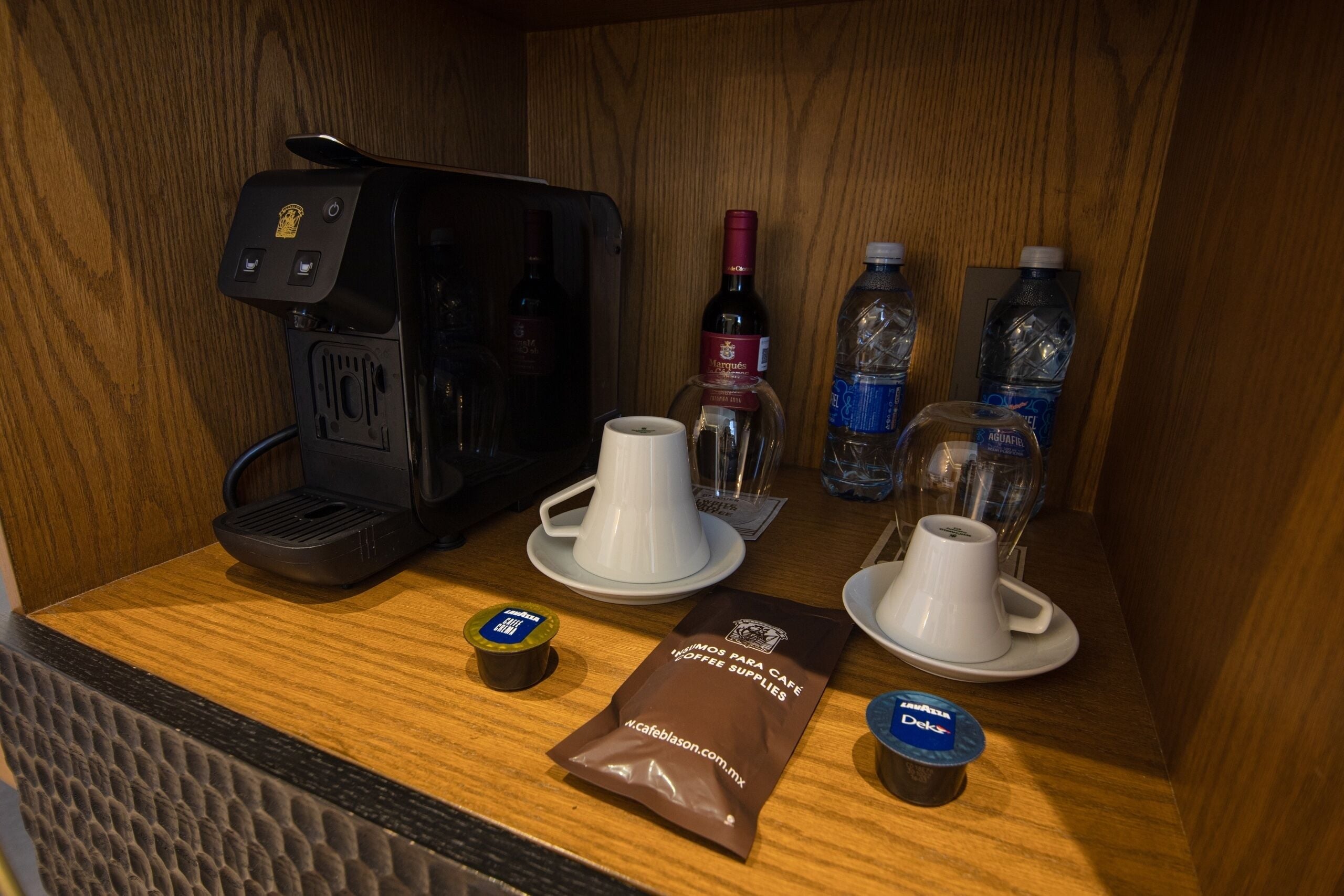 A coffee station with a coffee maker, cups, coffee pods, bottled water, and a bottle of wine.