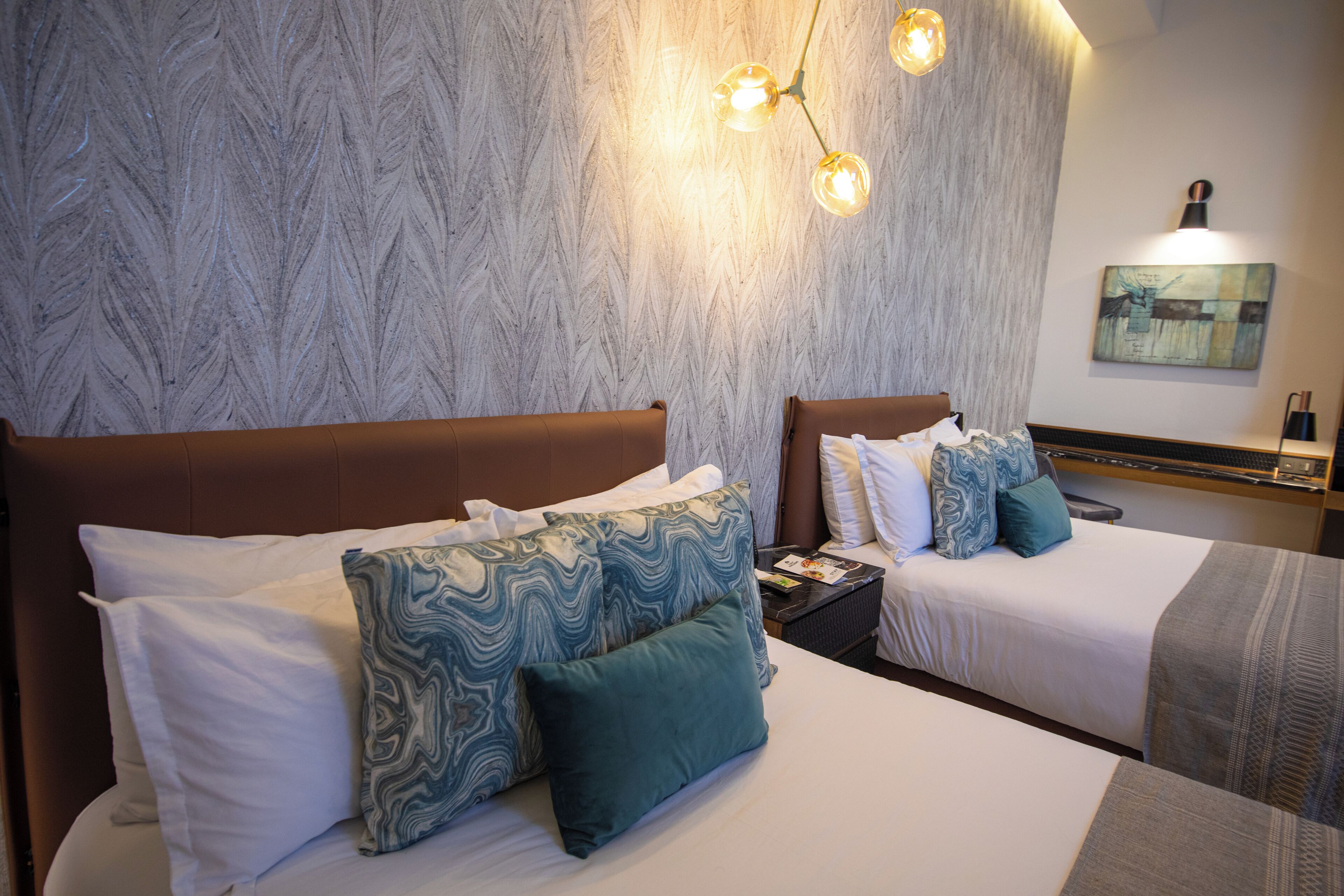 Een hotelkamer met twee bedden, elk voorzien van witte lakens en decoratieve kussens. Er is een nachtkastje met een lamp en een kleine tafel met een boek. De kamer heeft een structuurwand en een schilderij boven de bedden.