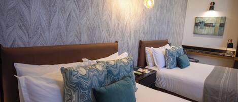 Een hotelkamer met twee bedden, elk voorzien van witte lakens en decoratieve kussens. Er is een nachtkastje met een lamp en een kleine tafel met een boek. De kamer heeft een structuurwand en een schilderij boven de bedden.