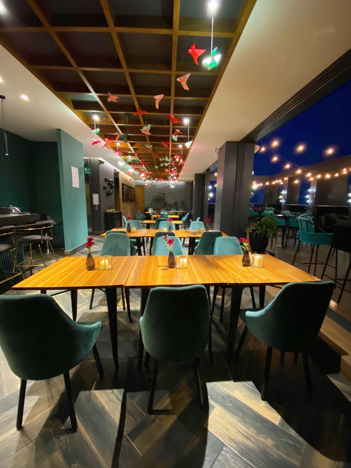 A restaurant with wooden tables and teal chairs, string lights, and hanging paper birds.