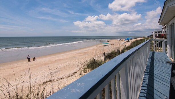 Uitzicht op een strand vanaf een balkon met houten hekwerk, met zicht op een zandstrand en enkele personen en strandparasols.