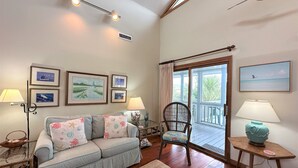 A living room with a sofa, a chair, a table with a lamp, framed pictures on the wall, and a view through a glass door. - Second Row Classic w/ Screened Porch; Across the Street From Beach Access; WiFi & Cable TV's (Edisto Beach)