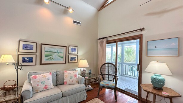A living room with a sofa, a chair, a table with a lamp, framed pictures on the wall, and a view through a glass door.