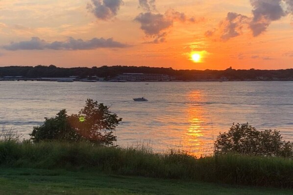 A sunset over a calm body of water with a boat and distant buildings. - Prime Lakefront East Okoboji Condo at Bridges Bay Jet Ski Lift rental available! (Arnolds Park)