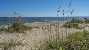 A beach with sand and sparse vegetation, a clear sky, and calm water.