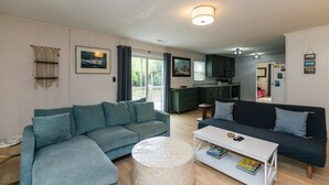 A living room with a sectional sofa, a round coffee table, and a kitchen in the background. - Villa Kunterbunt | Classic Beach Walk Cottage | Screened Porch & Sun Deck | Sleeps 8 | Pet Friendly (Edisto Beach)