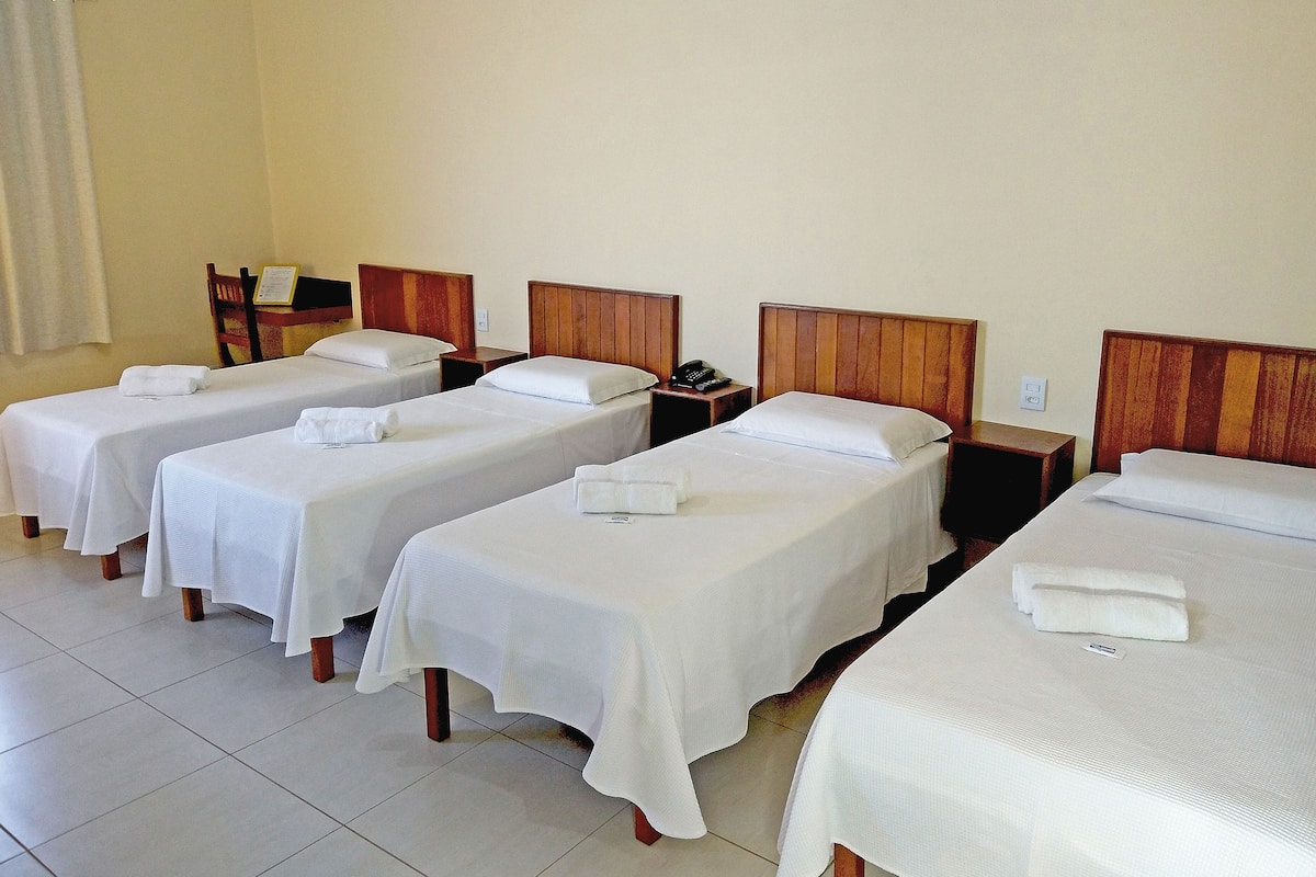Une chambre d’hôtel avec quatre lits simples, chacun doté d’une tête de lit en bois, de draps blancs et d’une table de chevet avec une lampe.