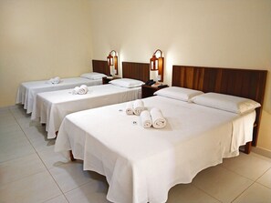 A hotel room with three beds, each with white linens and rolled towels. There are bedside tables with lamps and a telephone.