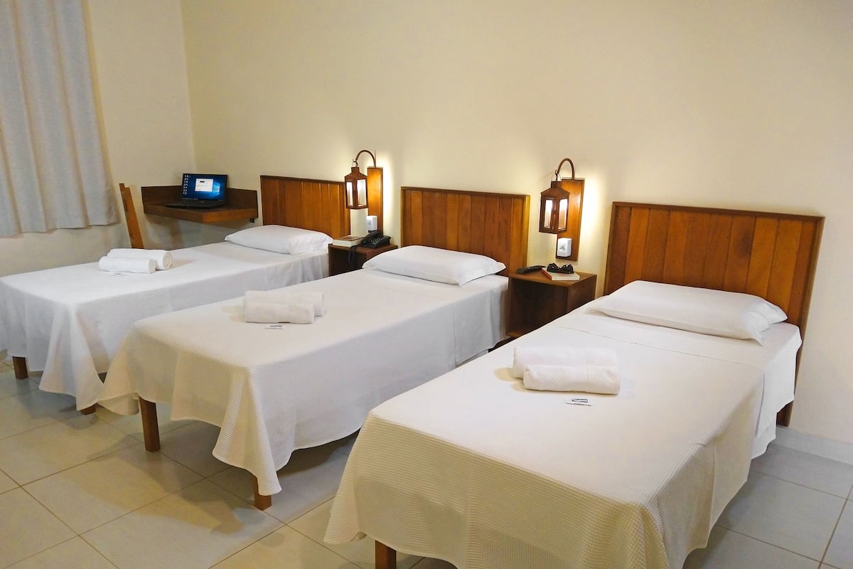 Une chambre d’hôtel avec trois lits simples, chacun recouvert d’une courtepointe blanche et agrémenté d’une serviette roulée. Il y a des tables de chevet avec des lampes et un petit écran affichant un message.