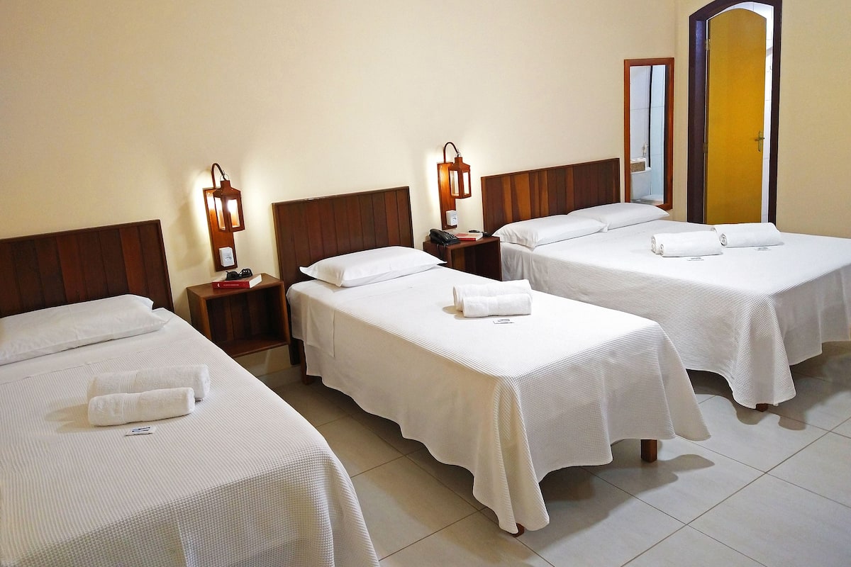 Une chambre d’hôtel avec trois lits simples, chacun équipé de draps et de serviettes blancs, de têtes de lit en bois et de lampes de chevet.