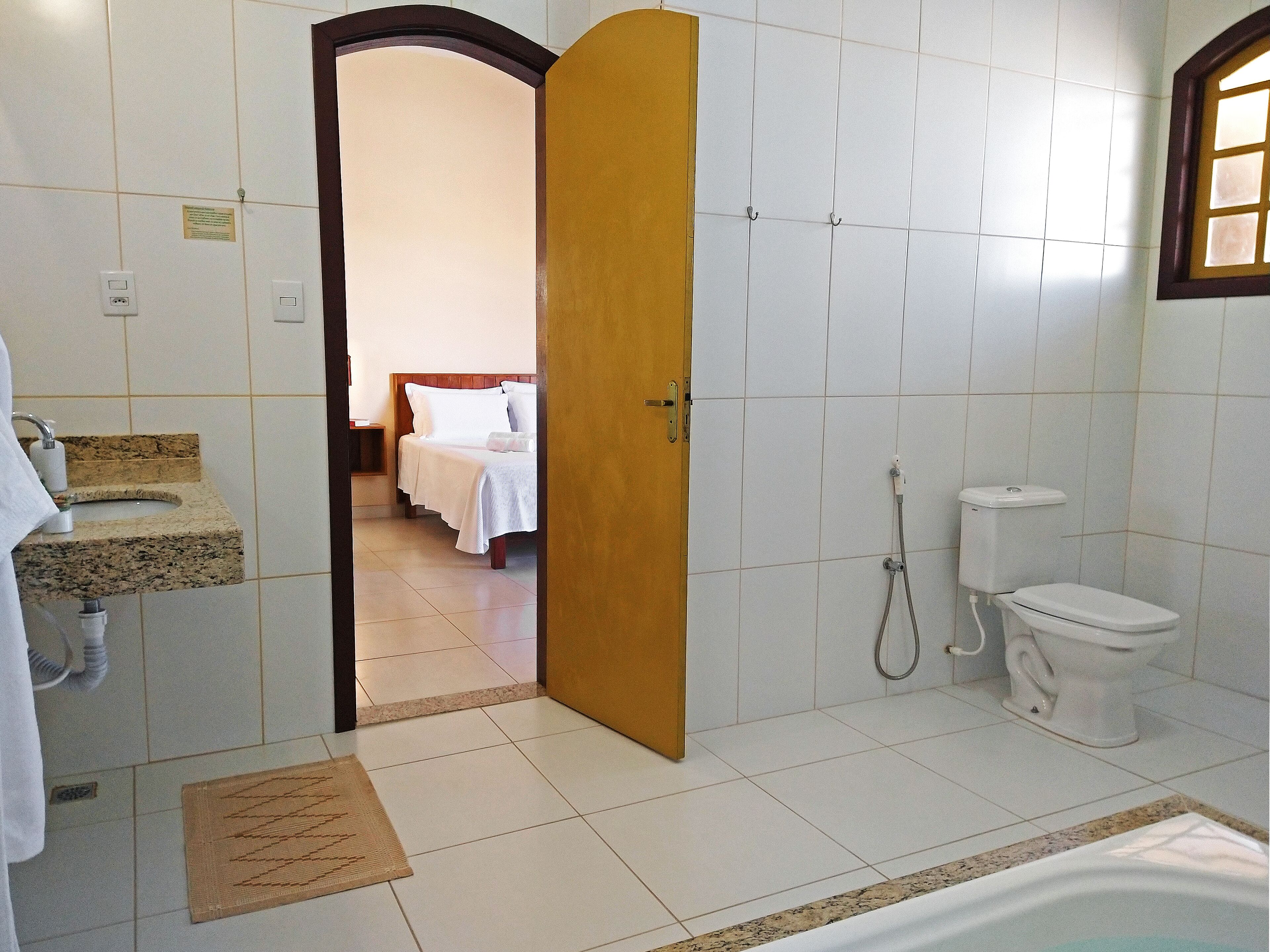 Un baño con lavamanos, inodoro, ducha y una cama visible a través de una puerta abierta.