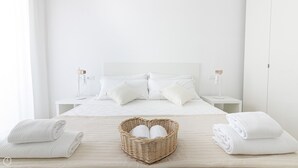 A minimalist bedroom with a large bed, white bedding, two bedside tables with lamps, and a heart-shaped basket with towels. - Rivazzurra Homes - 36 (Termoli)