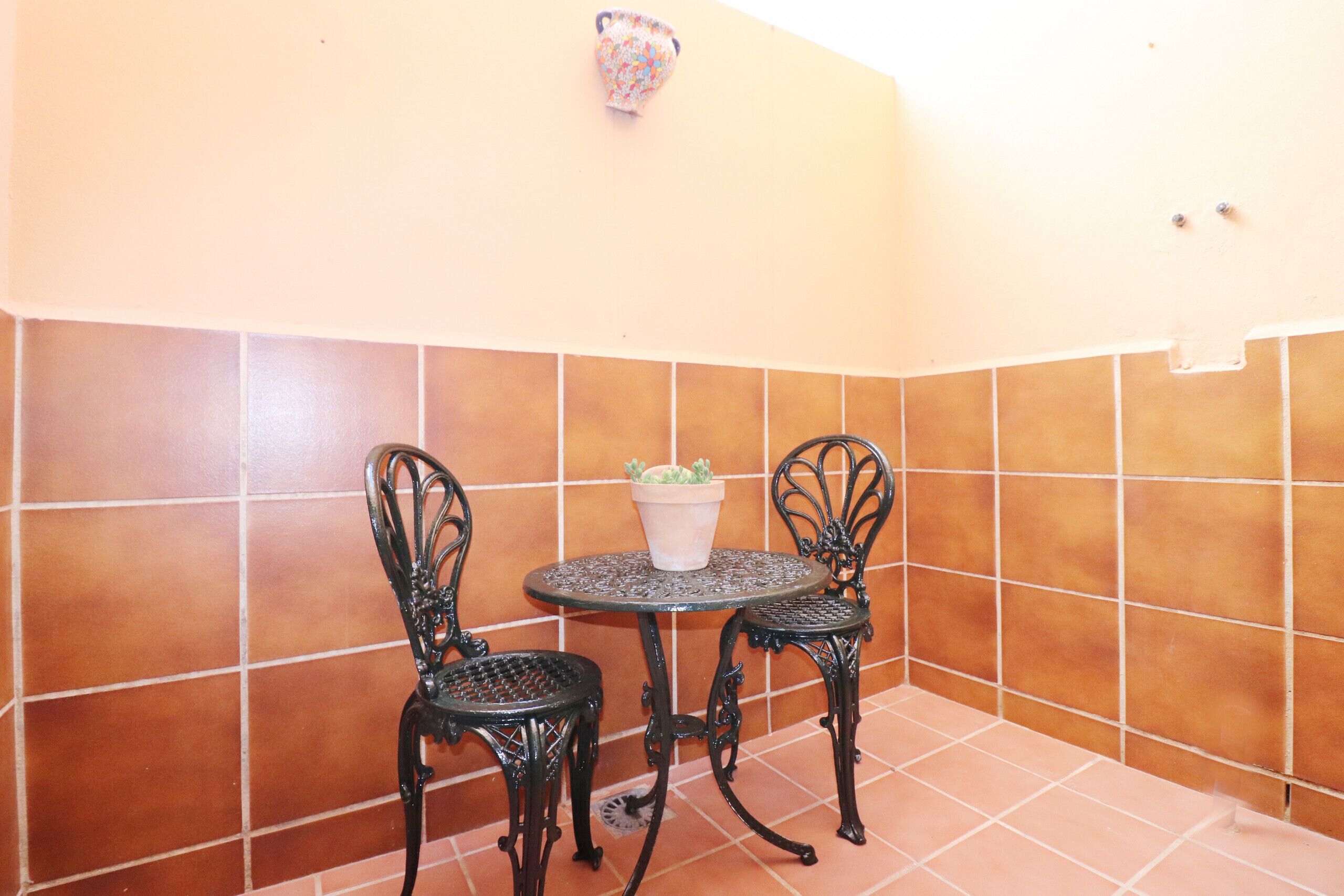 Un pequeño patio con dos sillas de metal y una mesa, una planta en maceta y una pared de azulejos.