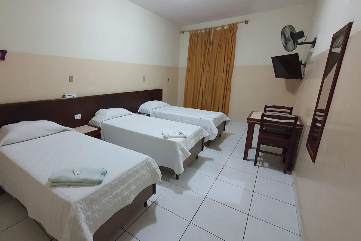 Une chambre d’hôtel avec trois lits simples, une chaise en bois, une petite table, un téléviseur et un ventilateur.