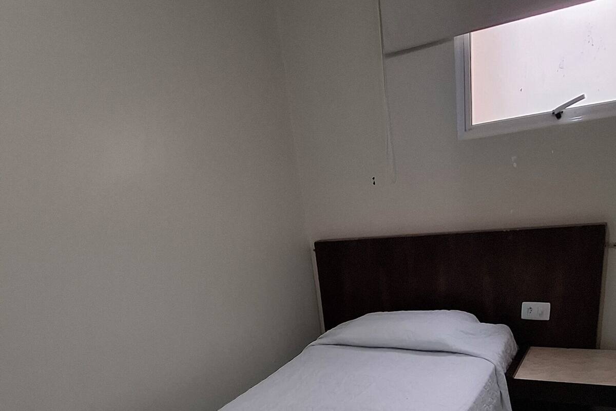 Une petite chambre avec un lit simple, recouverte d’une courtepointe blanche unie, dotée d’une tête de lit en bois et d’une fenêtre donnant sur le ciel.