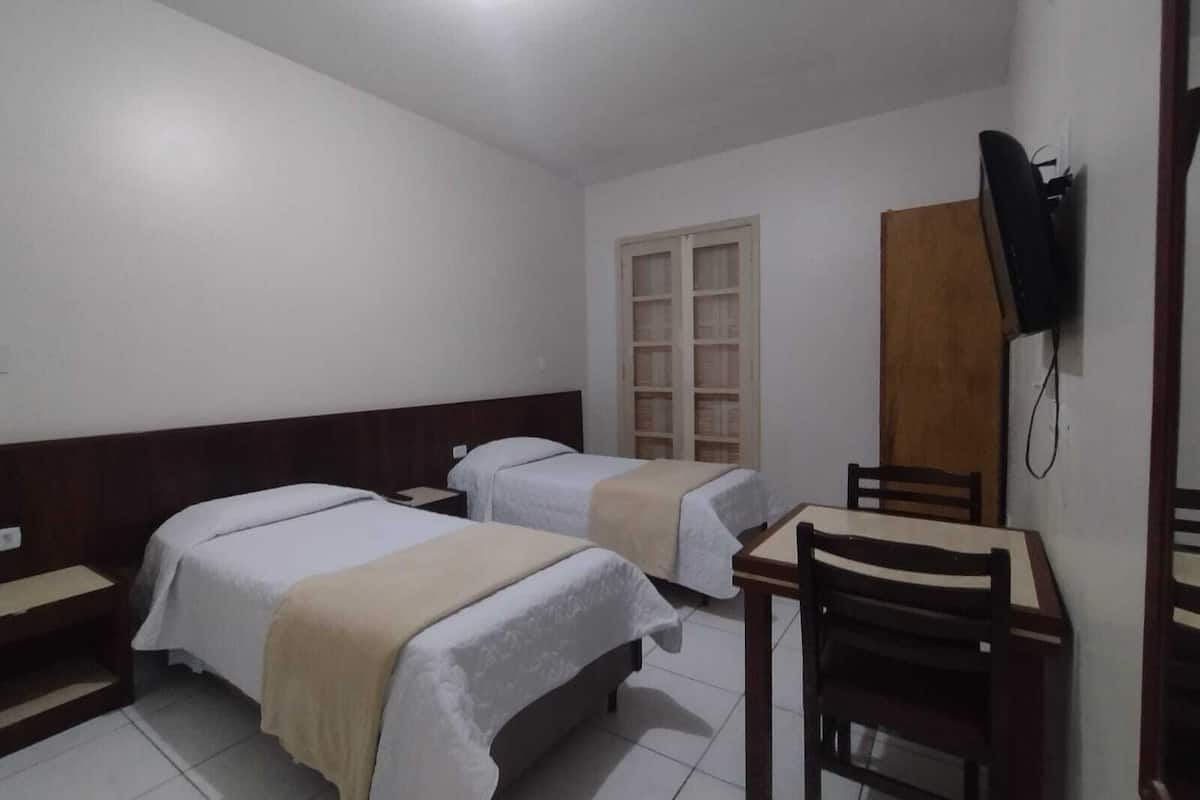 Une chambre d’hôtel avec deux lits, un bureau, une chaise, une armoire et un téléviseur fixé au mur.