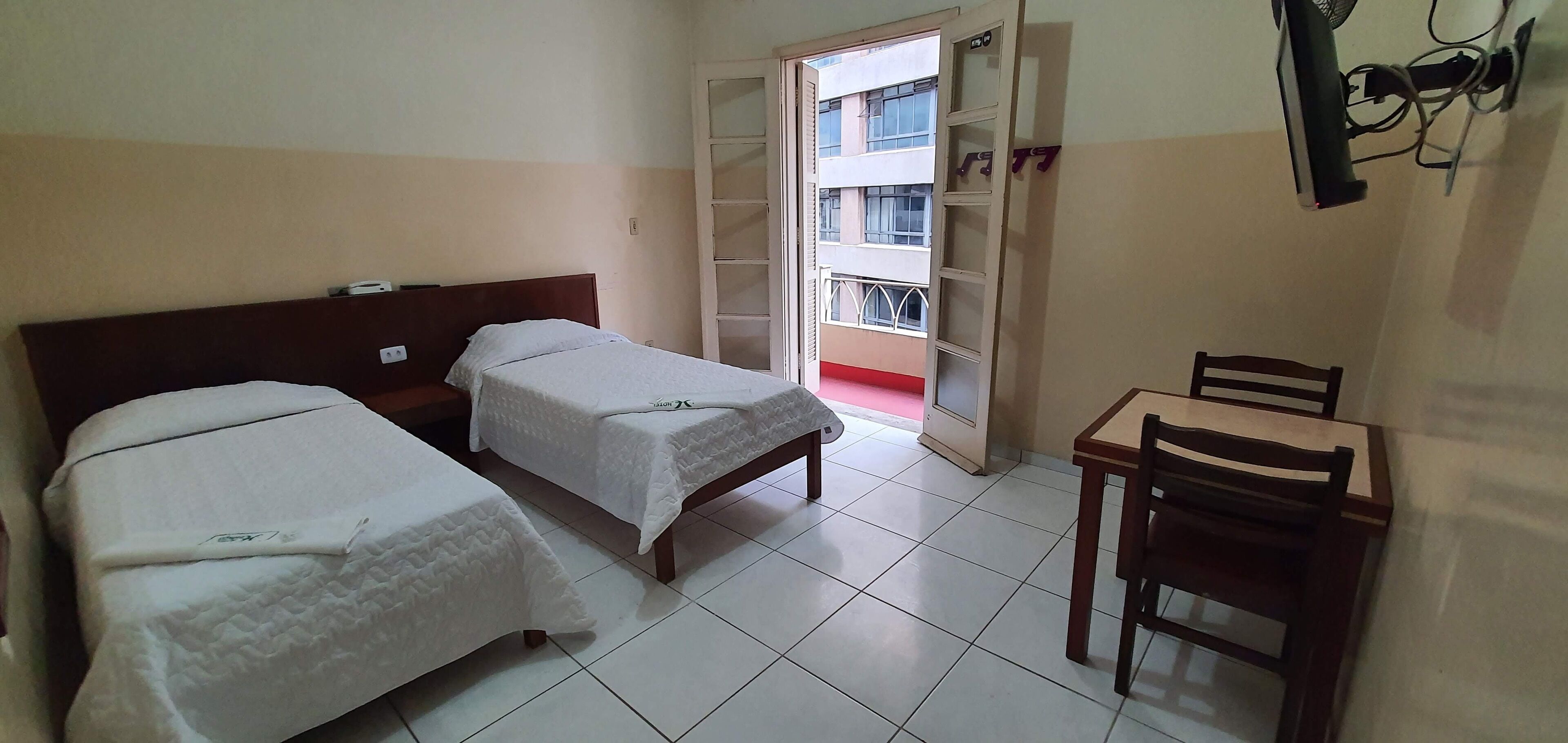 A hotel room with two single beds, a wooden desk with a chair, and a window with a view of a building.