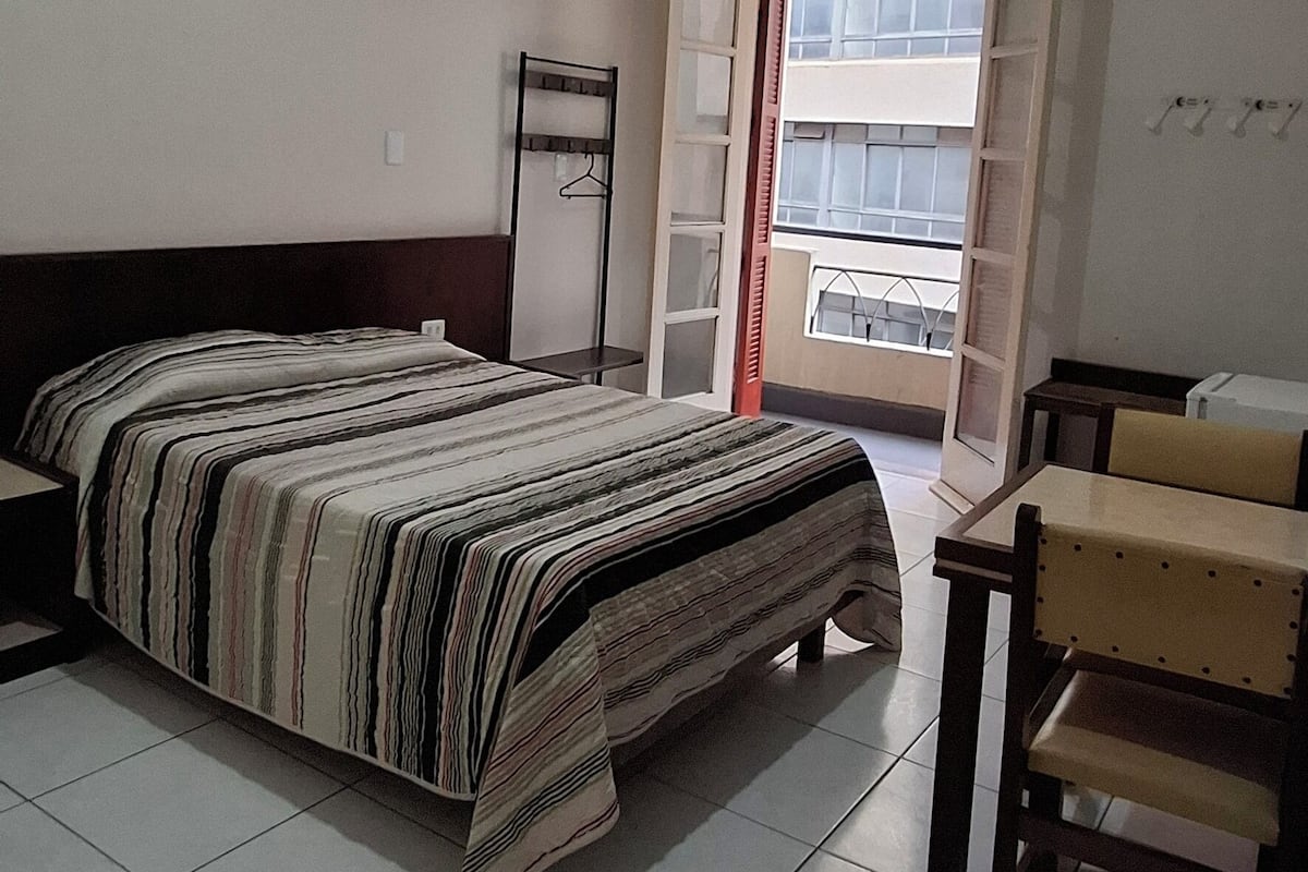 Une chambre à coucher comprenant un lit, un bureau, une chaise, un luminaire suspendu et une fenêtre avec des stores.